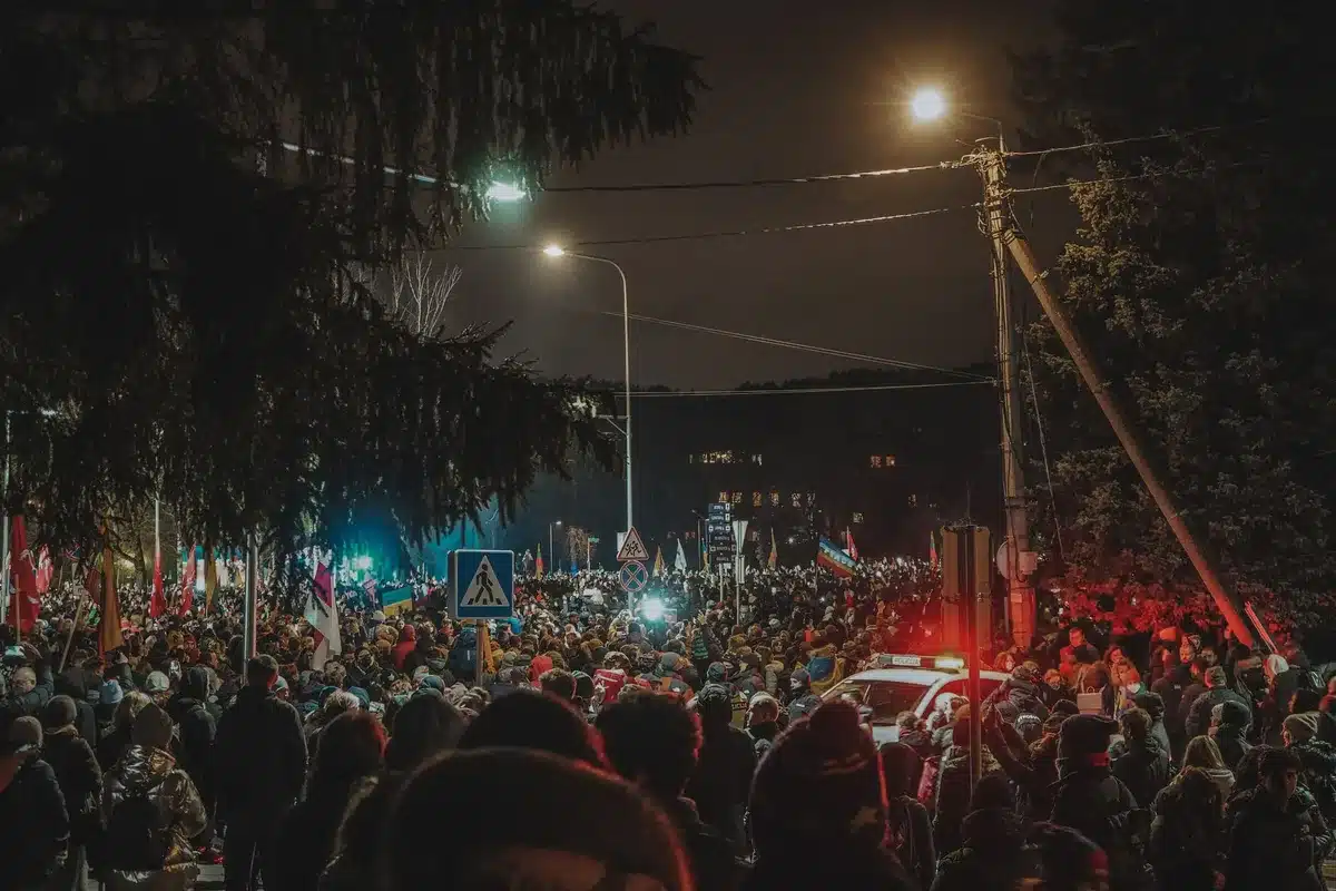 A political rally or protest scene with crowds and speakers on stage, flags and banners visible, daytime outdoor setting, political mobilization evident