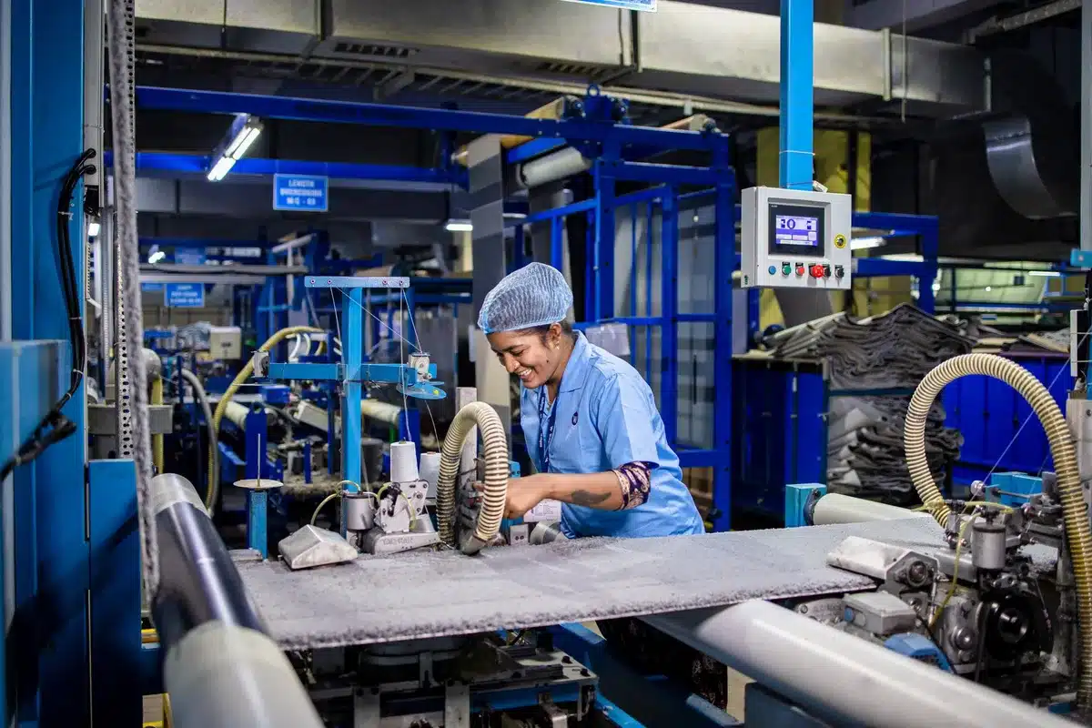 A drone assembly line with technicians working on unmanned systems at workstations, components arranged in organized rows. Hands adjust delicate electronics on small frames. Overhead lighting illuminates the precision manufacturing environment.