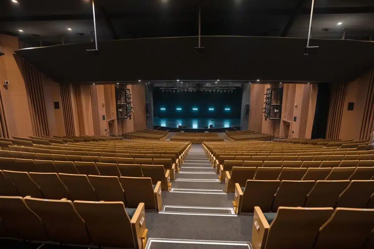 A university auditorium stage with empty rows of seats, a single podium under a spotlight, harsh shadows across the venue, ambient tension in the empty space