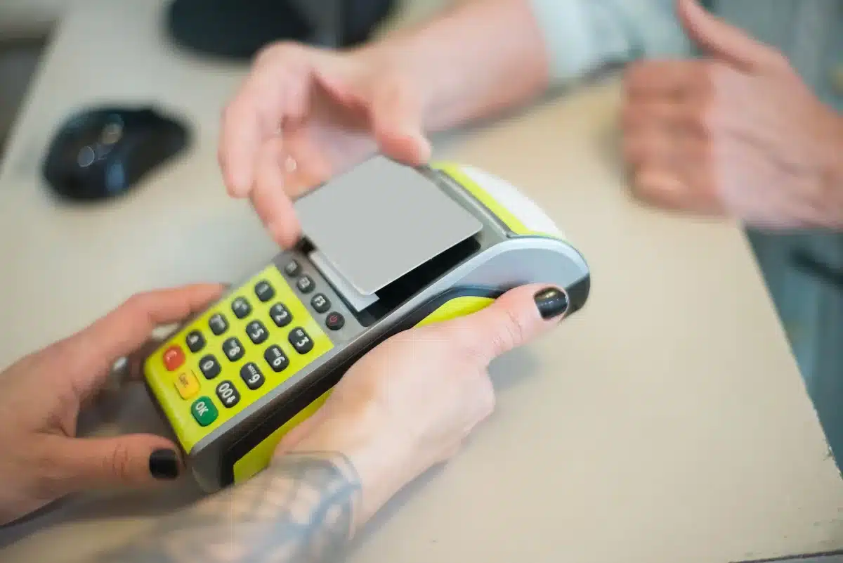 Retail checkout counter with multiple payment terminals visible, one displaying the tap-to-pay symbol, a few anonymous customers in soft focus in the background, modern store lighting