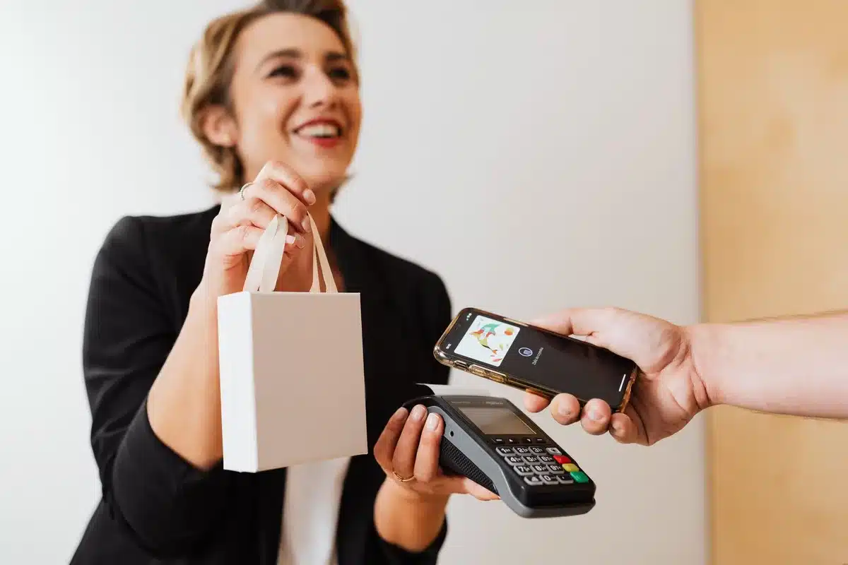 A retail checkout counter with multiple payment terminal options visible, a customer's hand reaching toward a modern NFC-enabled payment device, and store shelves blurred in the background