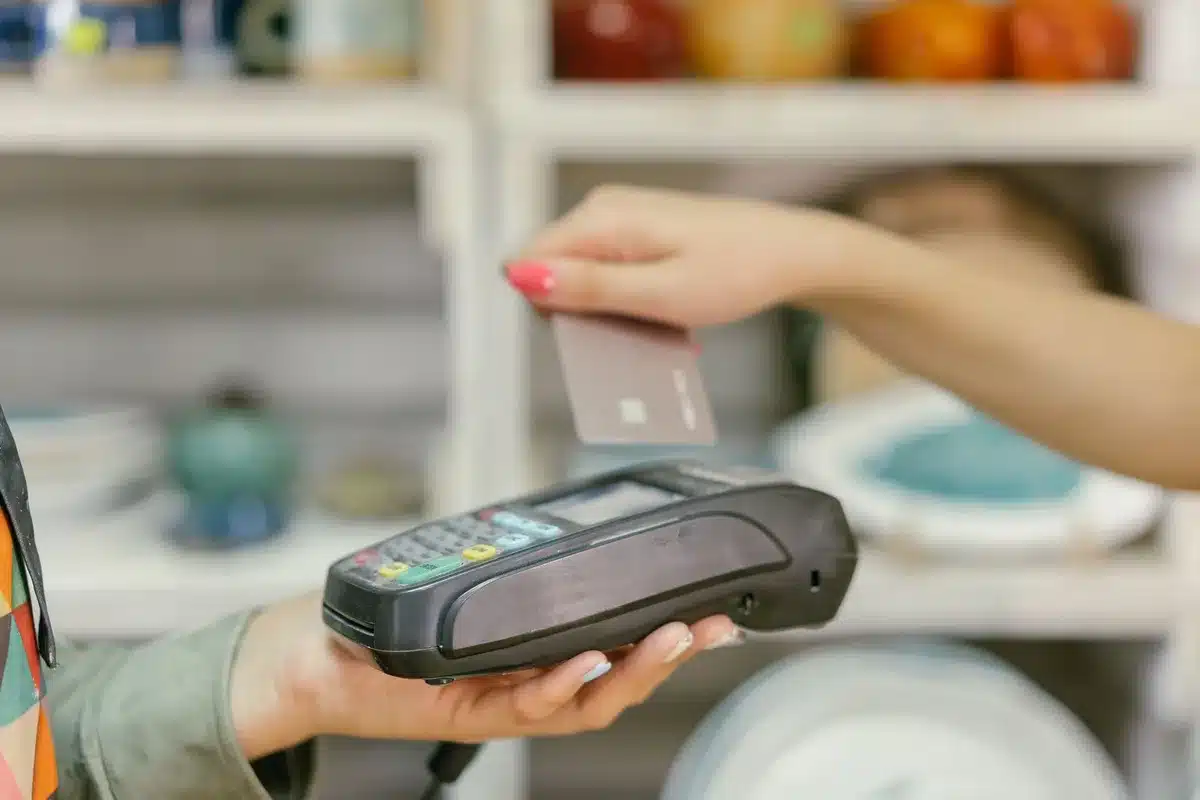 A retail checkout aisle with multiple payment terminals visible, one displaying the tap-to-pay symbol, a few anonymous customers in soft focus in the background, modern store lighting