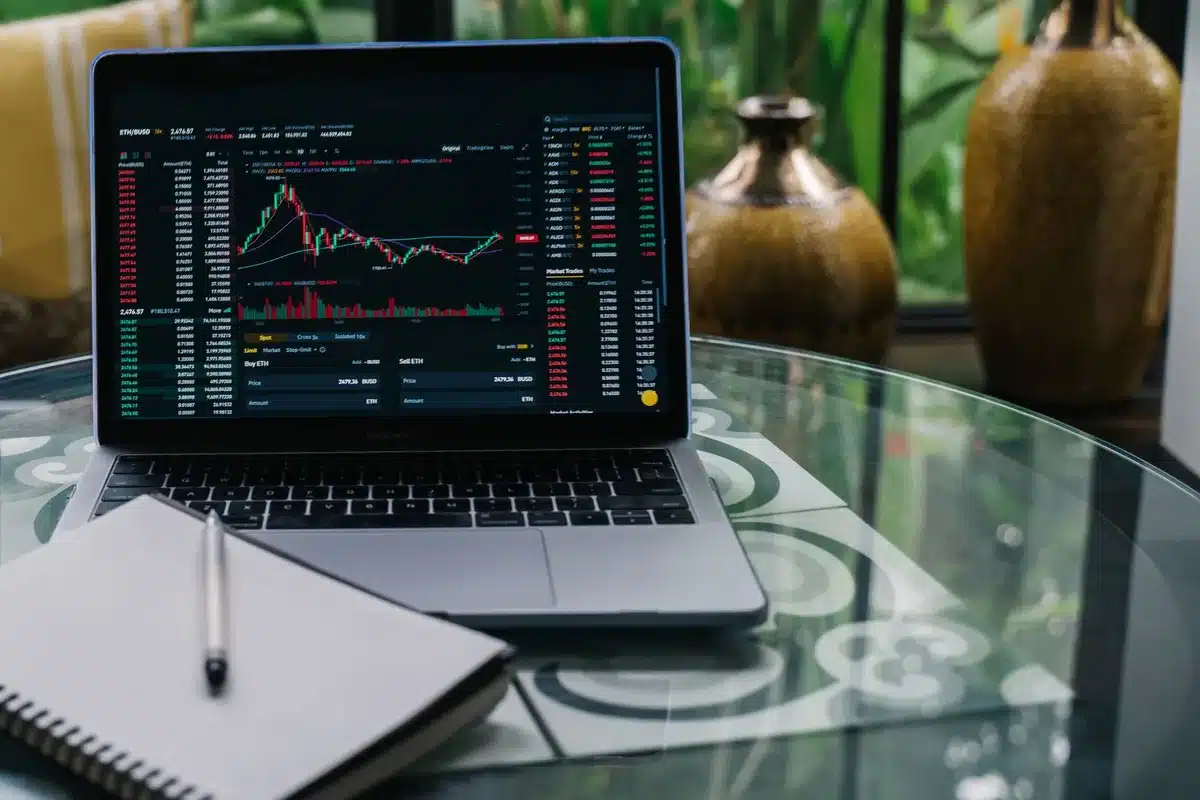 Stock market ticker display showing rising numbers in green, candlestick charts climbing upward, a trader's hand pointing at the screen