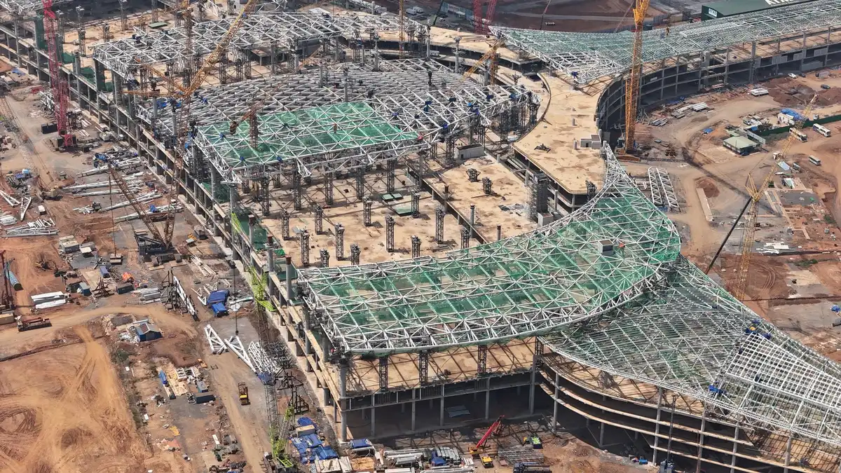 A construction site with steel framework rising against a clear sky, multiple cranes positioned above a large-scale building frame, safety barriers and equipment scattered across the foundation, dust and activity visible across the sprawling work area