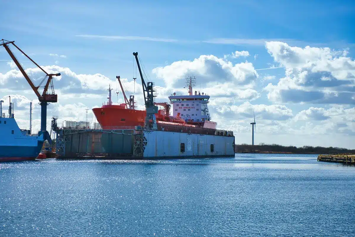 A cargo ship with visible damage, hull markings showing impact marks, deck equipment scattered, crew members working on repairs in daylight, aftermath