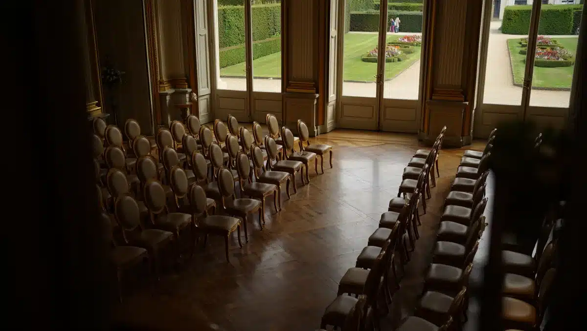 An empty, elegantly appointed formal room with tall windows overlooking manicured gardens, rows of empty chairs arranged for an official ceremony, soft evening light filtering through the windows, and a sense of anticipation before a state event.