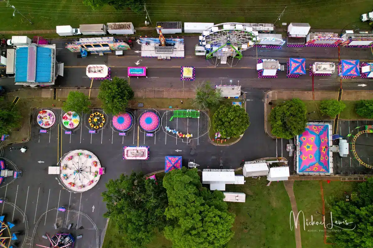 A crowd of fairgoers walking through the midway with colorful rides and food vendors, the massive wheel dominating the skyline behind them, capturing the energy and scale of opening day.