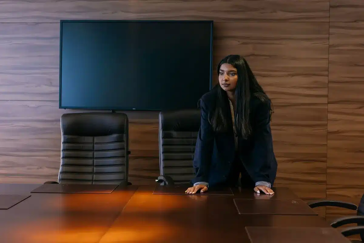 A formal boardroom table with five empty chairs positioned around it, one at the head, soft natural light filtering through tall windows overlooking a city skyline, a leather portfolio and pen on the table surface, subtle corporate aesthetic conveying governance and oversight