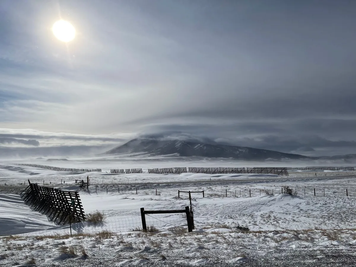A Wyoming rancher in work attire standing on open rangeland with distant mountains, weathered fence posts and grassland stretching to the horizon, embodying rural stewardship and land management.