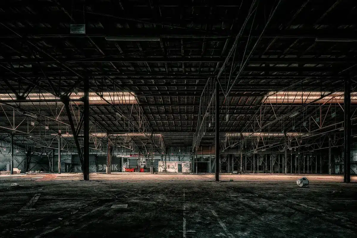 An empty industrial factory floor with shuttered windows and faded signage, dust particles in shafts of light, abandoned machinery silhouettes, economic decline atmosphere