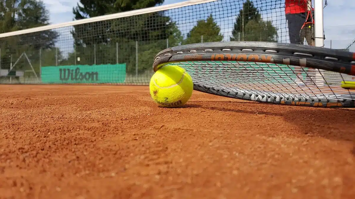 An anonymous close-up of a tennis racket leaning against a bench near an empty practice court, a folded towel and an unbranded water bottle beside it