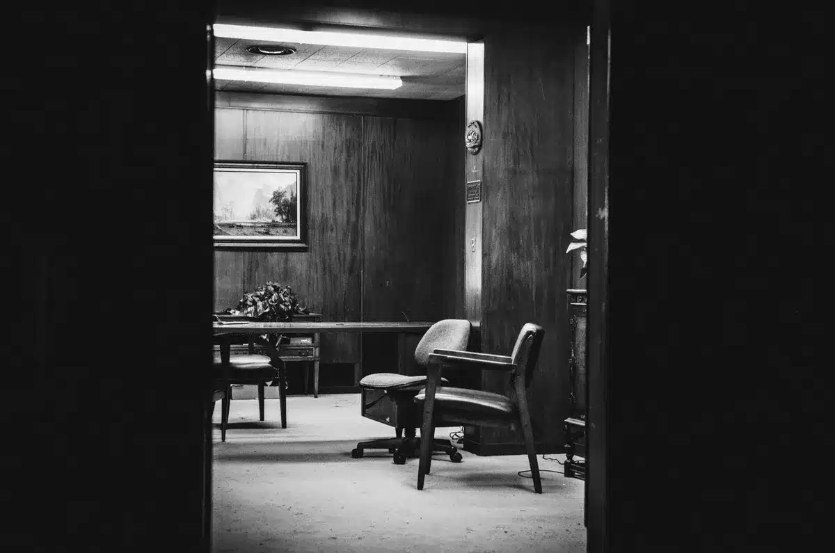 An empty, well-lit boardroom table with a single unoccupied leather chair pushed slightly back, a closed notebook and fountain pen on the table suggesting a recent departure