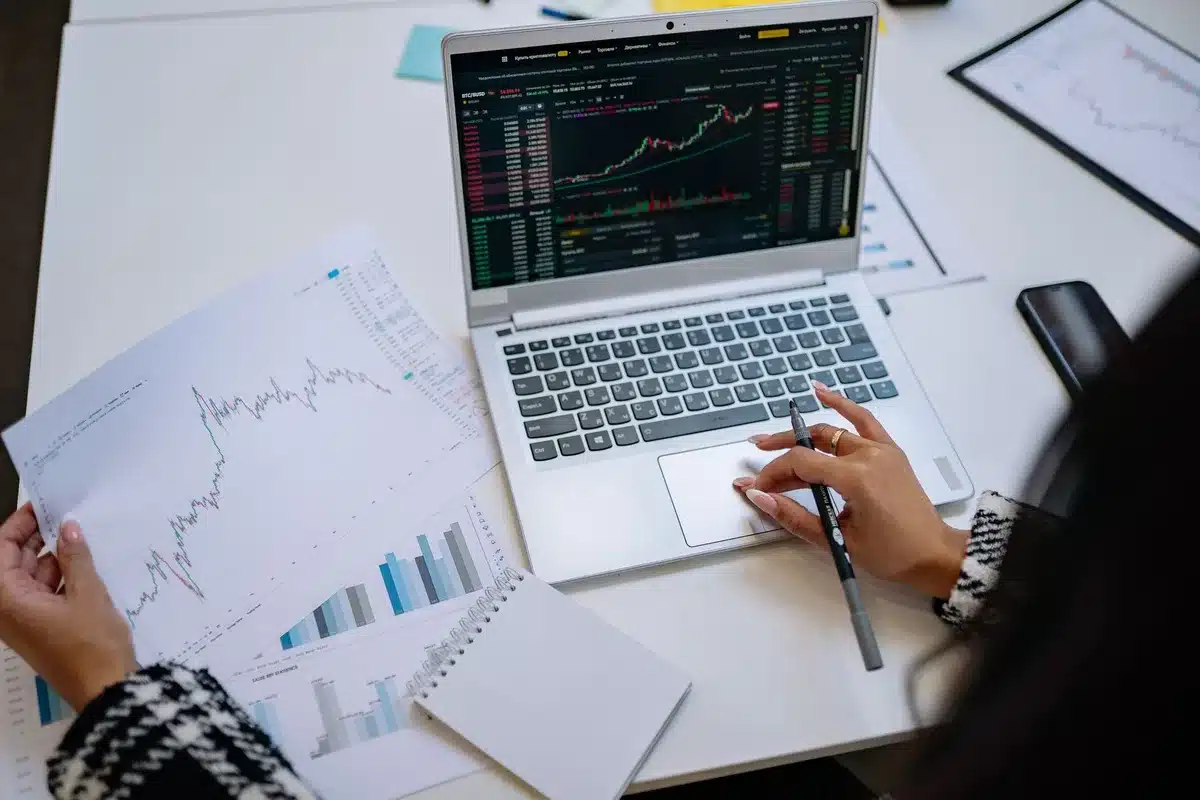 An anonymous trading floor screen showing price charts and a highlighted bitcoin price line around seventy-seven thousand dollars, with out-of-focus traders in the background