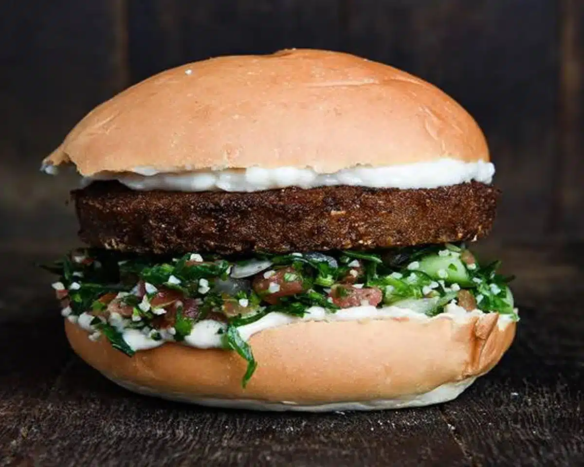 Clamshell box holding an anonymous, unbranded stacked burger with a taller sesame bun and visible fresh lettuce and tomato