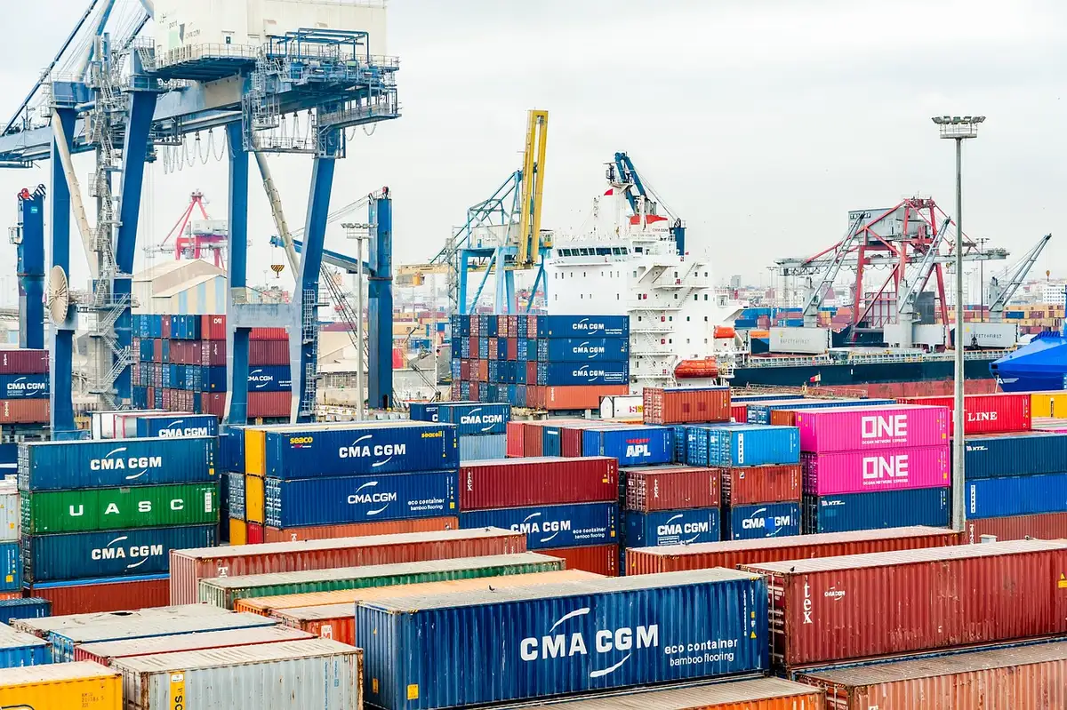 Shipping containers at a port or border crossing, tariff notices visible, industrial landscape with cranes