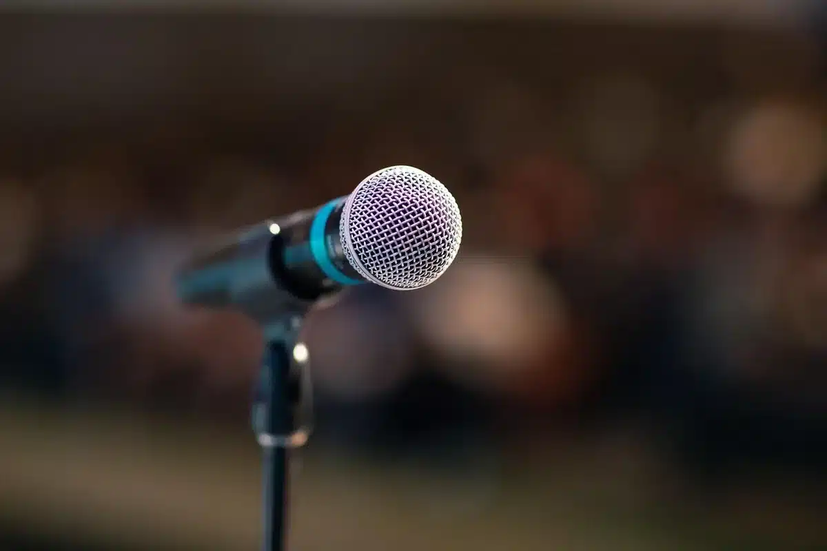 A young woman at a professional political event, speaking at a microphone with formal government building architecture visible in soft focus behind her, serious expression, professional attire