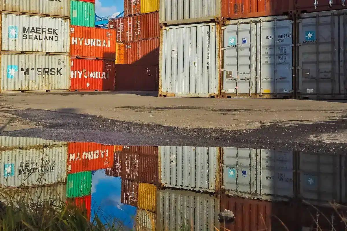 Stacks of shipping containers at a border port facility, early morning light, American and Canadian flags visible in soft focus, representing the flow of trade between the nations