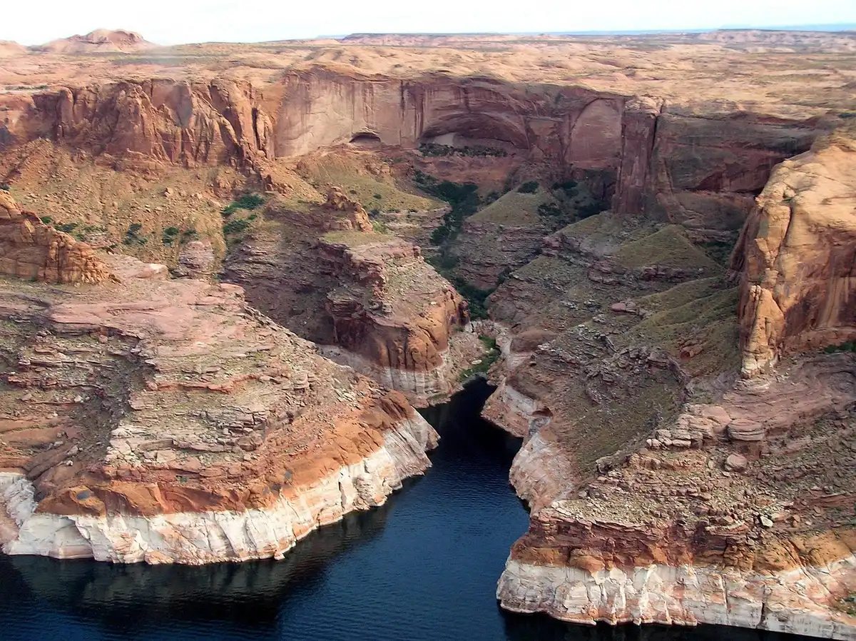 Aerial view of Glen Canyon Dam with dramatically reduced water levels upstream, showing the scale of Lake Powell's decline and the critical infrastructure at risk.