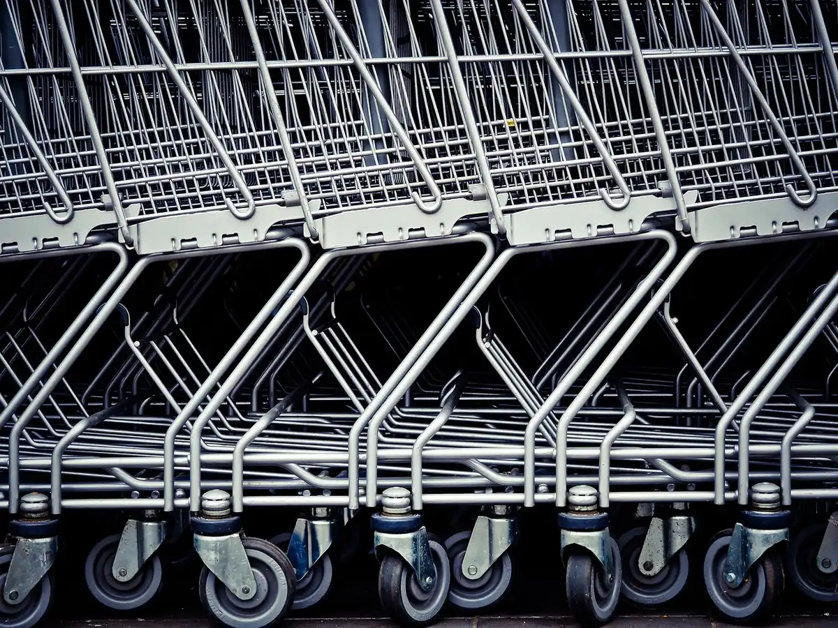 Anonymous shopper pushing a nearly full cart past supermarket shelves, price tags visible but unreadable, a slightly tense posture suggesting rising grocery costs