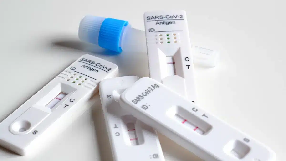 An anonymous close-up of a generic rapid test kit on a kitchen counter, an instruction leaflet blurred in the background, a hand reaching for the swab