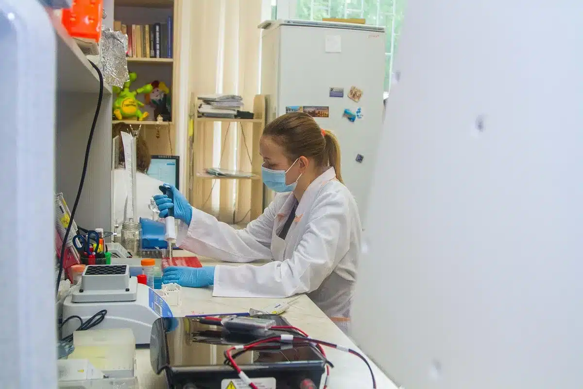 An anonymous laboratory bench showing a labelled sample tube beside a PCR machine, gloved hands not identifiable preparing a sample