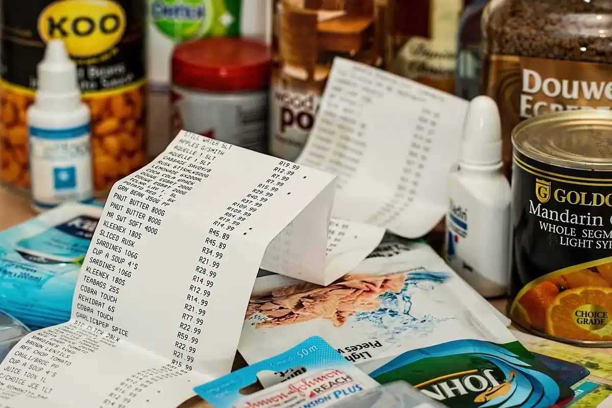 A grocery store shelf display of packaged ground beef in various trays and packages, fluorescent lights reflecting off the plastic wrapping, price tags visible at different heights, anonymous shoppers browsing in soft focus behind
