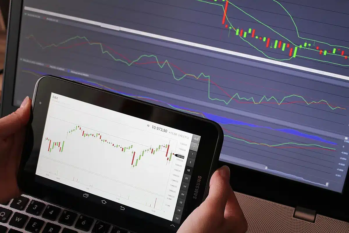 close-up of anonymous hands pointing at a tablet showing a candlestick FX chart, the tablet resting on a desk next to a notebook and a coffee cup