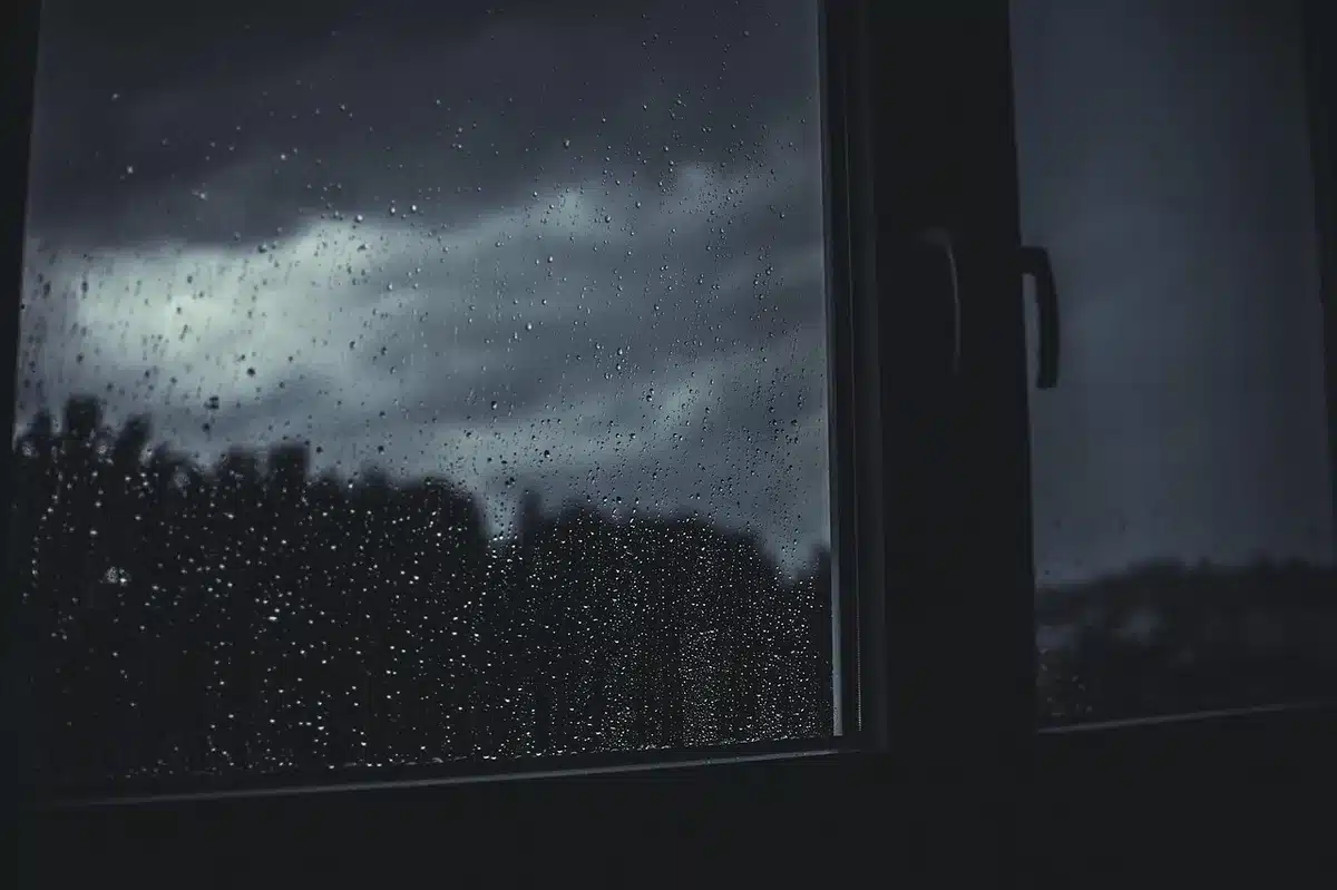 Darkened residential street with a few streetlights glowing through heavy rain and mist, a car's headlights barely visible through the downpour, utility lines sagging overhead