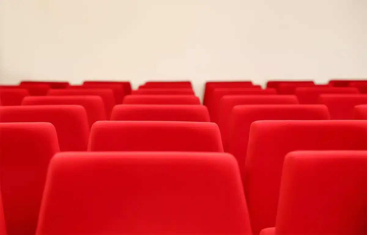 A dim conference stage with an empty lectern and a single spotlight, an anonymous audience in shadow watching large projection screens showing abstract AI code — tension