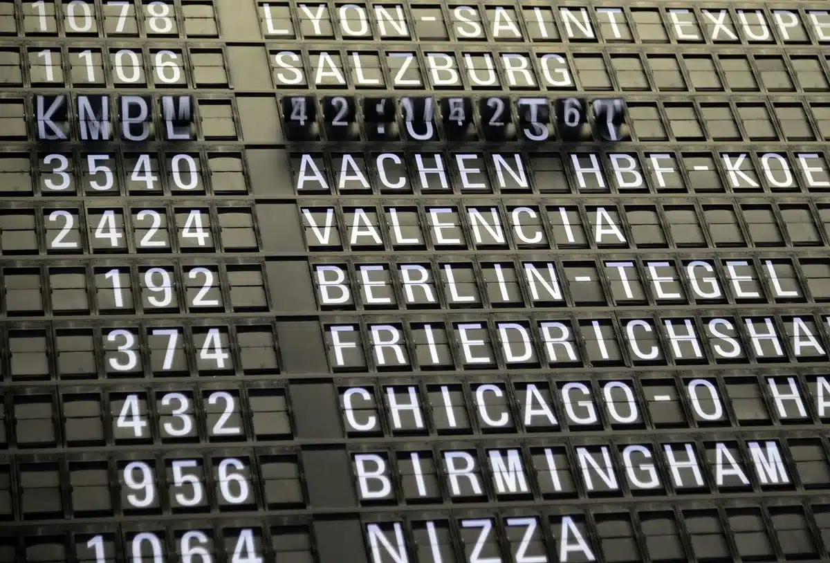 Inside an airport terminal with passengers checking flight information boards displaying cancellations and delays, expressions of concern visible, fluorescent lighting casting a clinical glow