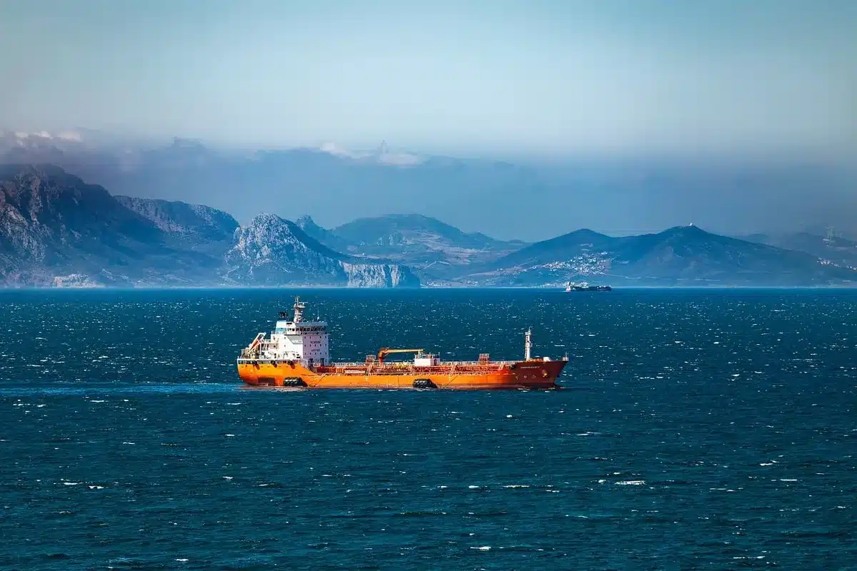 An oil tanker silhouetted against a sunset over calm waters, with faint smoke in the distance, evoking supply route vulnerability and geopolitical risk