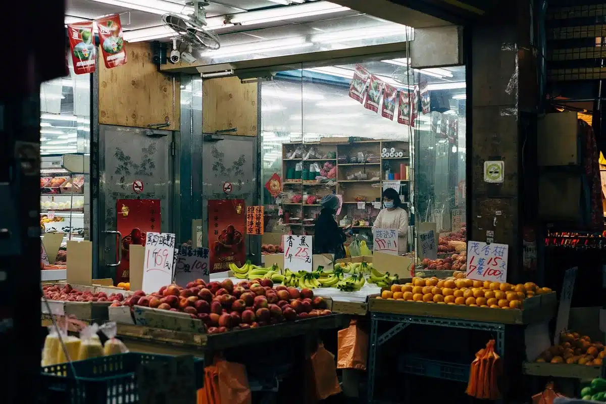 A grocery store produce section with colorful fresh fruits and vegetables arranged on tiered displays under bright store lighting, shoppers examining items up close, clean aisles and organized bins