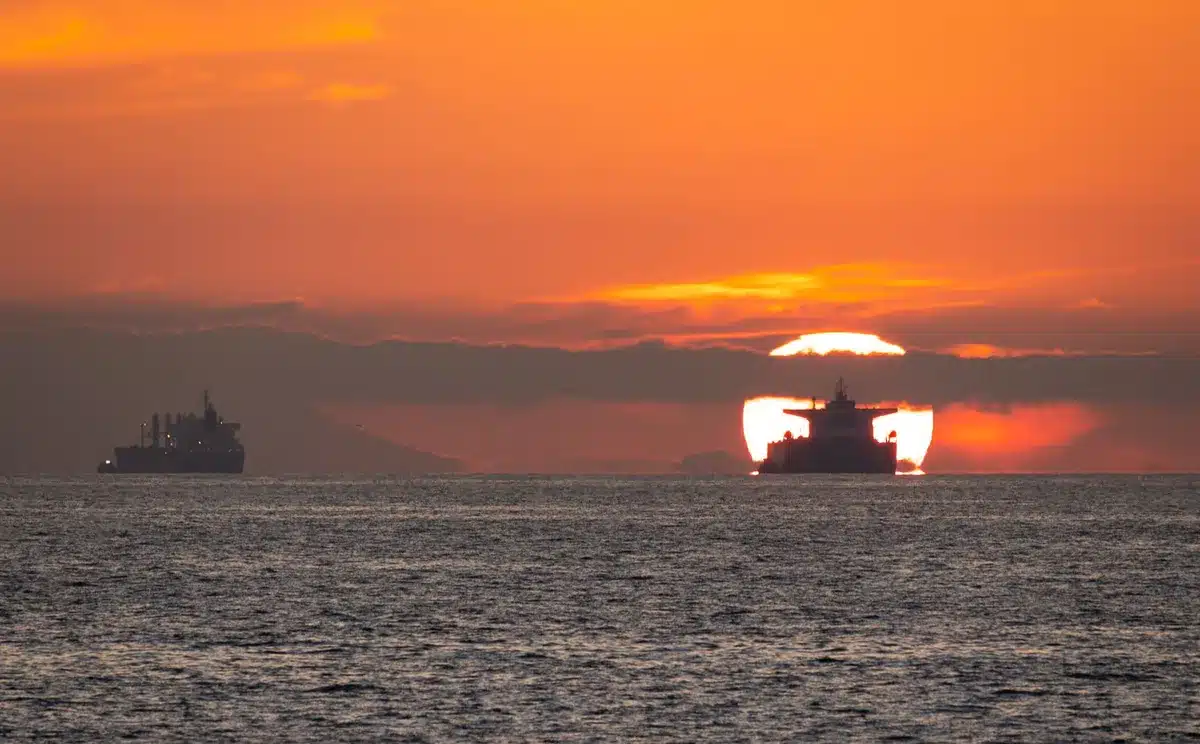 An empty stretch of busy shipping lane at dusk with several anonymous tankers silhouetted, low smoke rising from one vessel near the horizon — tension