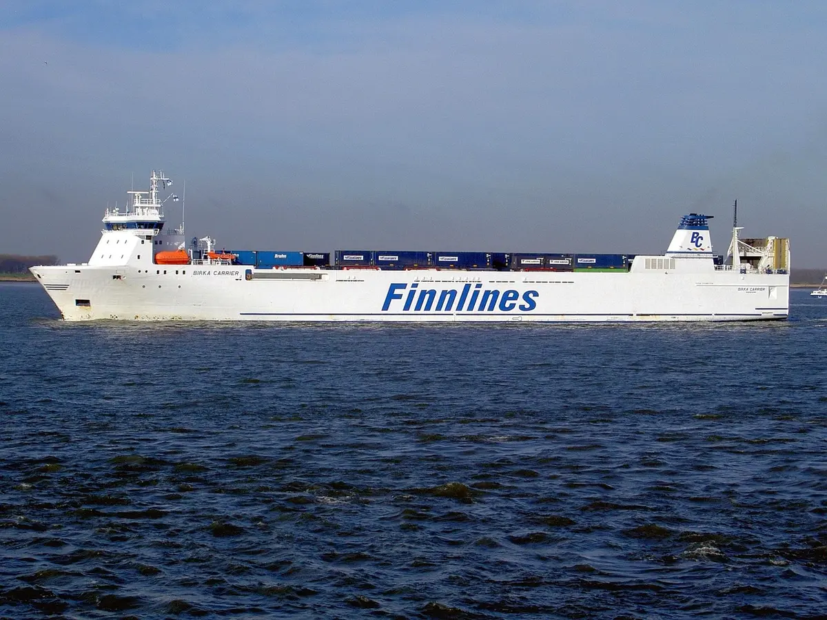 A dim, anonymous cargo vessel at dusk with a distant plume on the horizon; no logos or identifiable marks, empty deck and maritime safety buoys in foreground
