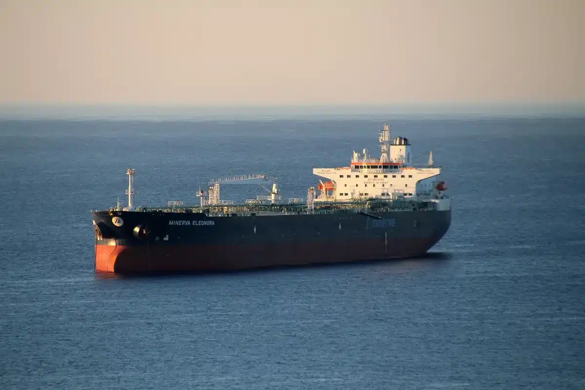 An empty shipping channel at dusk with several anonymous cargo tankers silhouetted under a hazy sky, one tugboat in the distance; no flags or logos visible