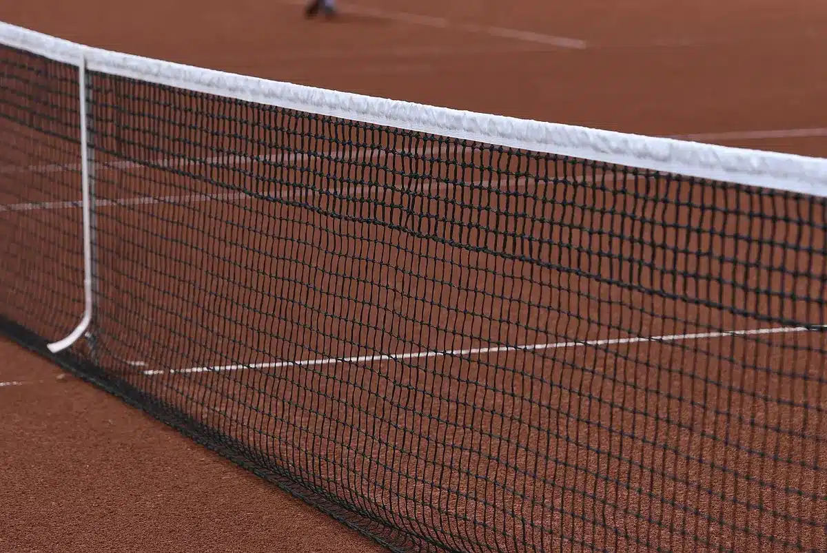 Empty tournament court at dusk with an unbranded tennis net and lone ball on the baseline; quiet stands in the background