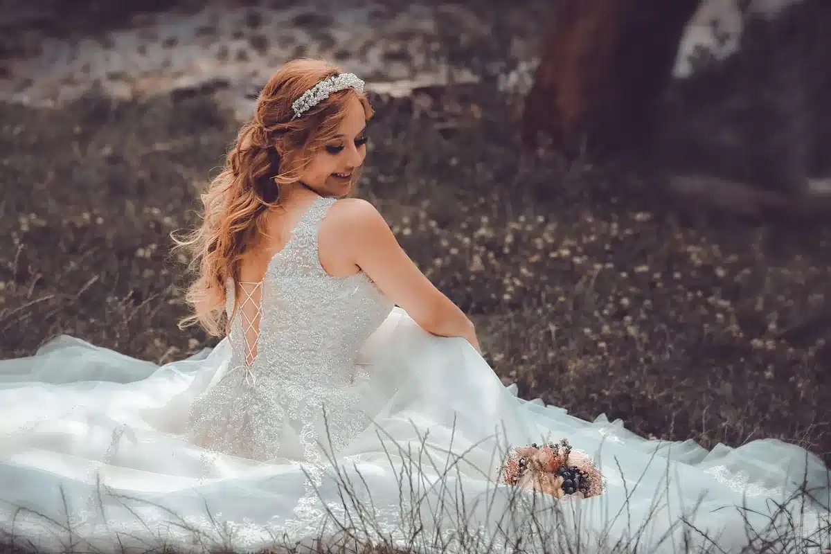 A single anonymous woman turned away from the camera in a softly lit bridal boutique, hands holding a white dress at her shoulders, empty chairs and blurred floral arrangements in the background