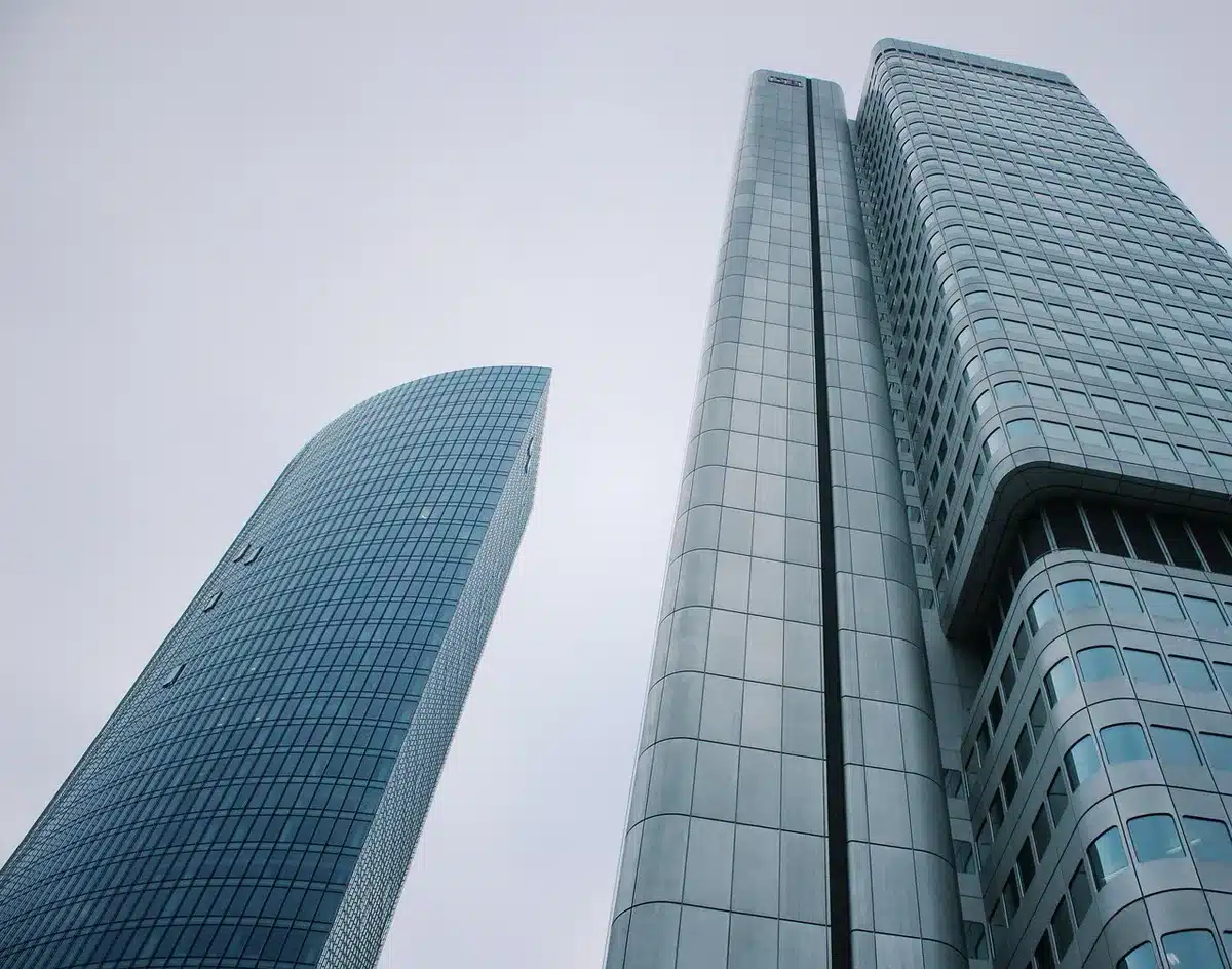 A modern office building exterior with glass windows reflecting daylight, a busy street with pedestrians and business professionals walking past, representing economic activity and workplace momentum