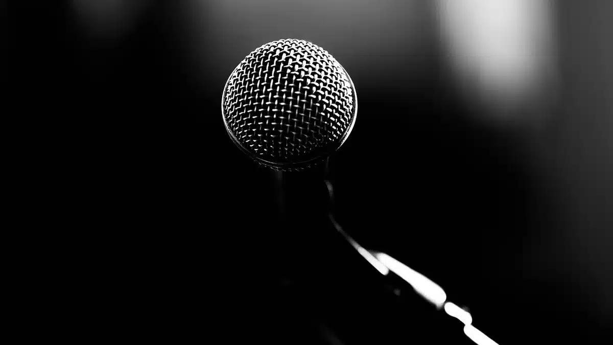 Empty studio stage with a single microphone on a stand under a pool of light, dust motes visible, empty chairs in the dark background, evocative of media coverage