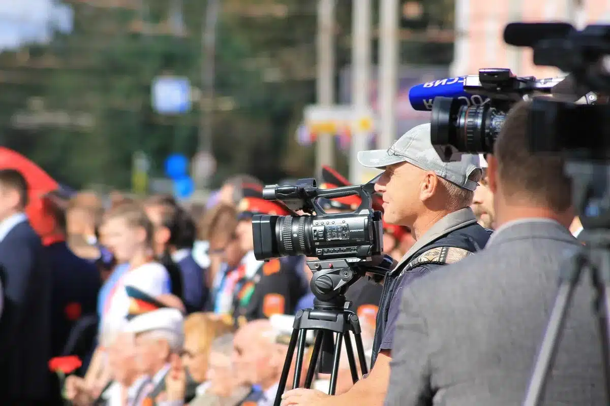 Anonymous figure at a podium approached by a cluster of microphones and distant photographers, empty nameplates on the table visible, capturing the tense press-line atmosphere