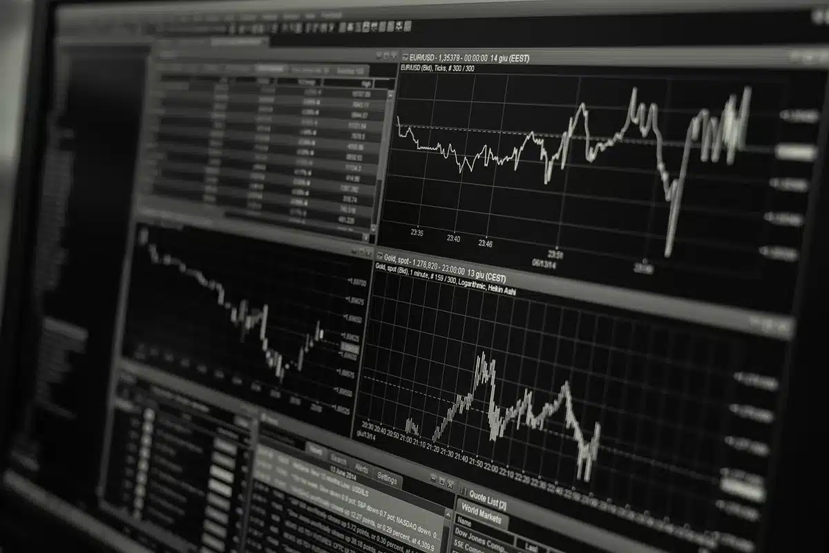Gold bars stacked on a trading desk, Bitcoin symbol glowing on a screen, financial charts with downward trending lines, hands holding precious metal coins