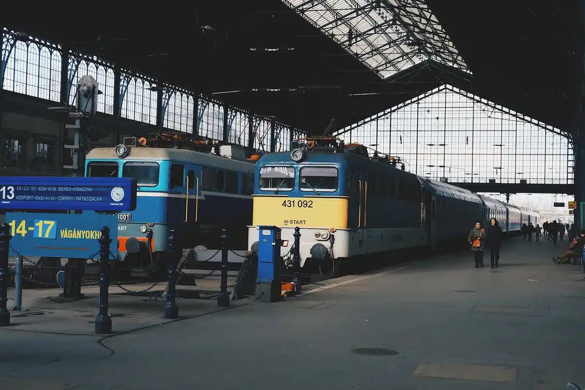 A diplomatic envoy descending train steps in a war-torn capital, armed security visible, the city skyline bearing marks of conflict in the background, daylight gray and tense