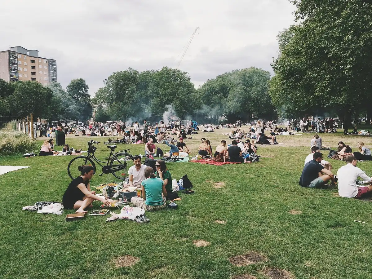 Diverse crowds gathered in a city park with picnic blankets, food vendors, and families relaxing on grass, with autumn-colored trees in the background.
