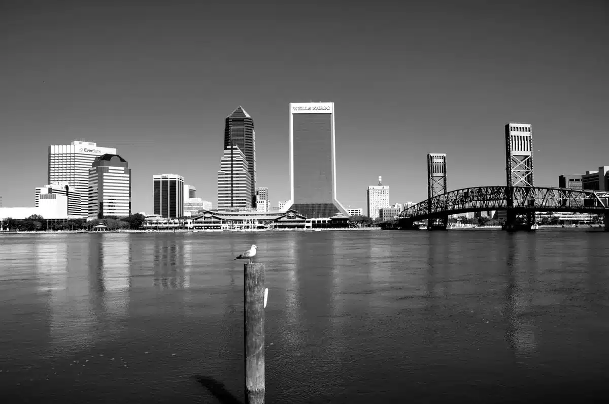 Two small riverfront towns seen from across a waterway at golden hour, storefronts and a low skyline, no identifiable signage or faces