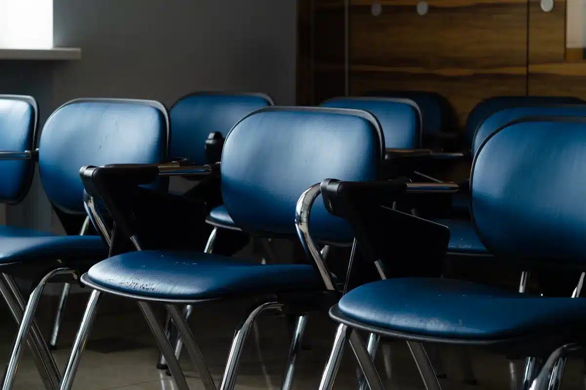 anonymous silhouette of a lone figure in an empty hearing room facing a bank of microphones and a long wood table — anticipatory, no real persons