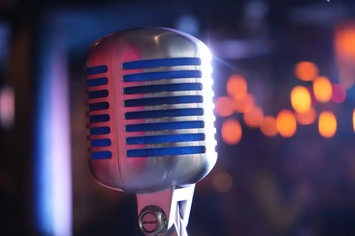 An empty staging area with an unbranded vintage microphone on a small round table under a single spotlight, a coiled cable trailing away into shadow, dust motes visible in the beam