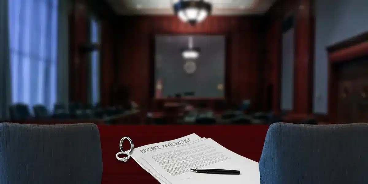 A dimly lit empty federal courtroom with a single lawyer's chair pushed back from the counsel table, scattered case files on the table and papers fanned open, conveying legal motion and unresolved questions