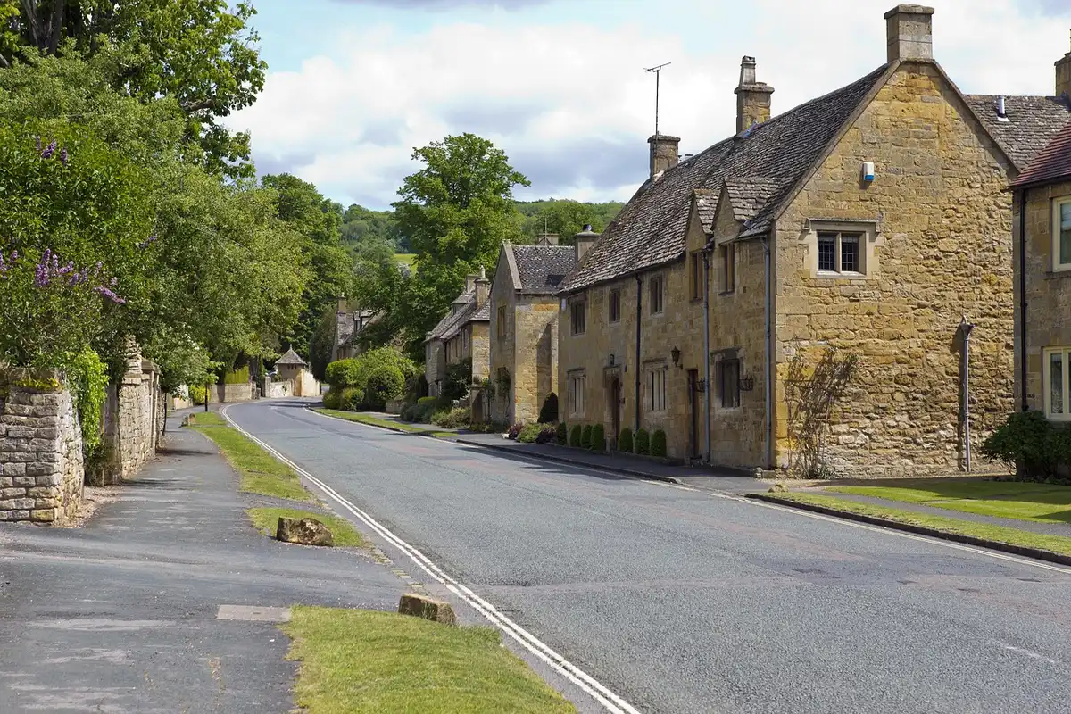 A winding tree-lined country lane in the English Cotswolds, golden stone cottages nestled among rolling green hills, autumn light filtering through mature oaks, a private drive disappearing into manicured grounds, peaceful rural seclusion
