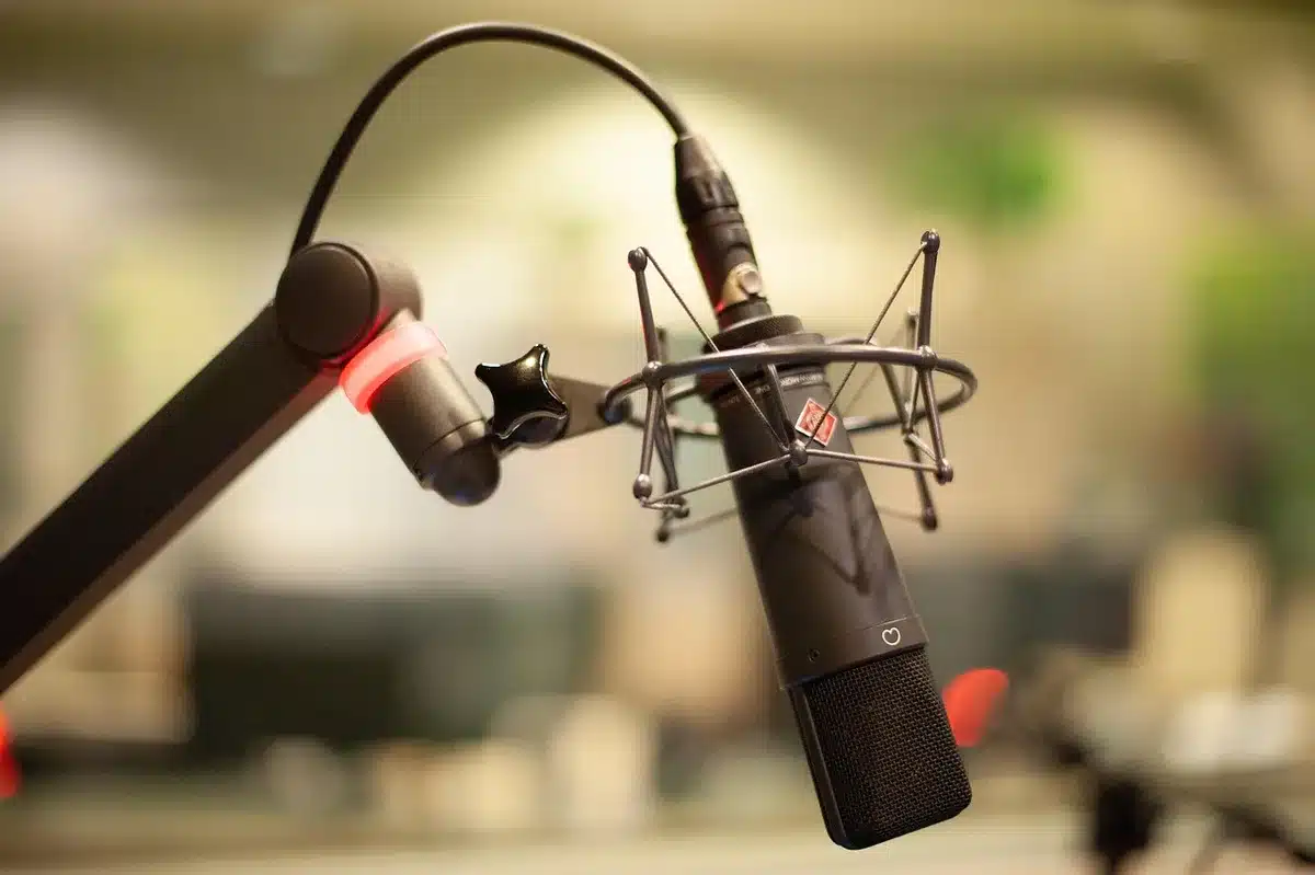 A small radio studio table with two anonymous microphones facing each other, an unbranded cassette recorder and folded show notes on the surface, a dim neon sign out of focus in the background