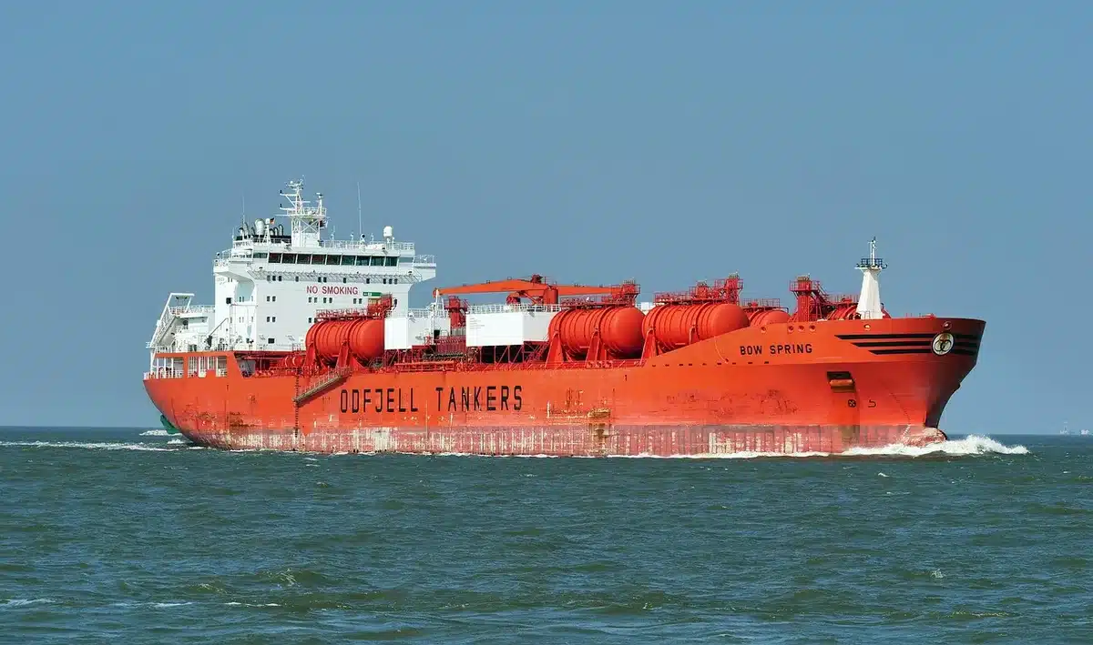 Tanker ship silhouetted against dawn light in a narrow waterway, with distant coastlines visible and radar domes on shore, representing the strategic maritime chokepoint under dispute